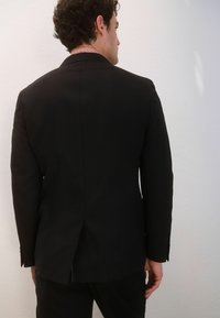 Jachetă de costum neagră personalizată, cu o deschidere simplă în spate, guler cu model contrastant vizibil. Textură netedă, formă structurată, mâneci lungi.