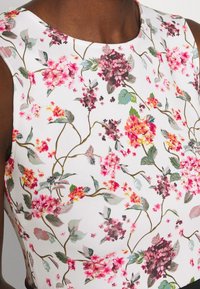 Gros plan d'un haut blanc sans manches avec un motif floral rose, violet et orange et des feuilles vertes sur une peau foncée.