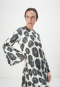 Faltenkleid in Weiß mit großem grauem Blumenmuster, ausgestattet mit langen, ausladenden Ärmeln und einem runden Ausschnitt. Glattes, leichtes Material.