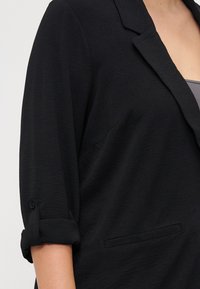 Blazer noir en tissu texturé, avec des manches retroussées trois quarts, un col ouvert et des poches latérales.