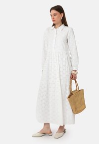 White cotton long dress with a button-up collar, tiered design, eyelet texture, and subtle dot pattern. Paired with a woven basket bag.