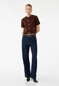 Braunes, kurzes Strickoberteil mit kurzen Ärmeln und goldenen Knöpfen, kombiniert mit dunkelblauen, geraden Jeans und schwarzen, spitzen Absatzschuhen.