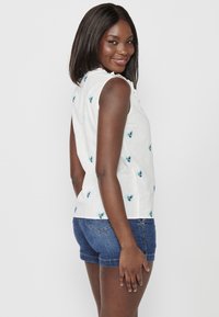 Weißes ärmelloses Top mit floralem Stickerei in blauen Tönen, Rüschensaum und lässiger Passform, kombiniert mit Jeansshorts.