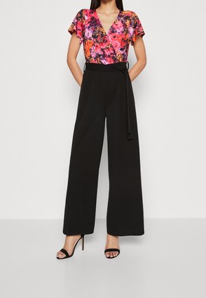 Femme portant un haut cache-cœur floral à manches courtes et un pantalon noir large taille haute avec ceinture à la taille, assortis à des sandales à talons aiguilles noires.