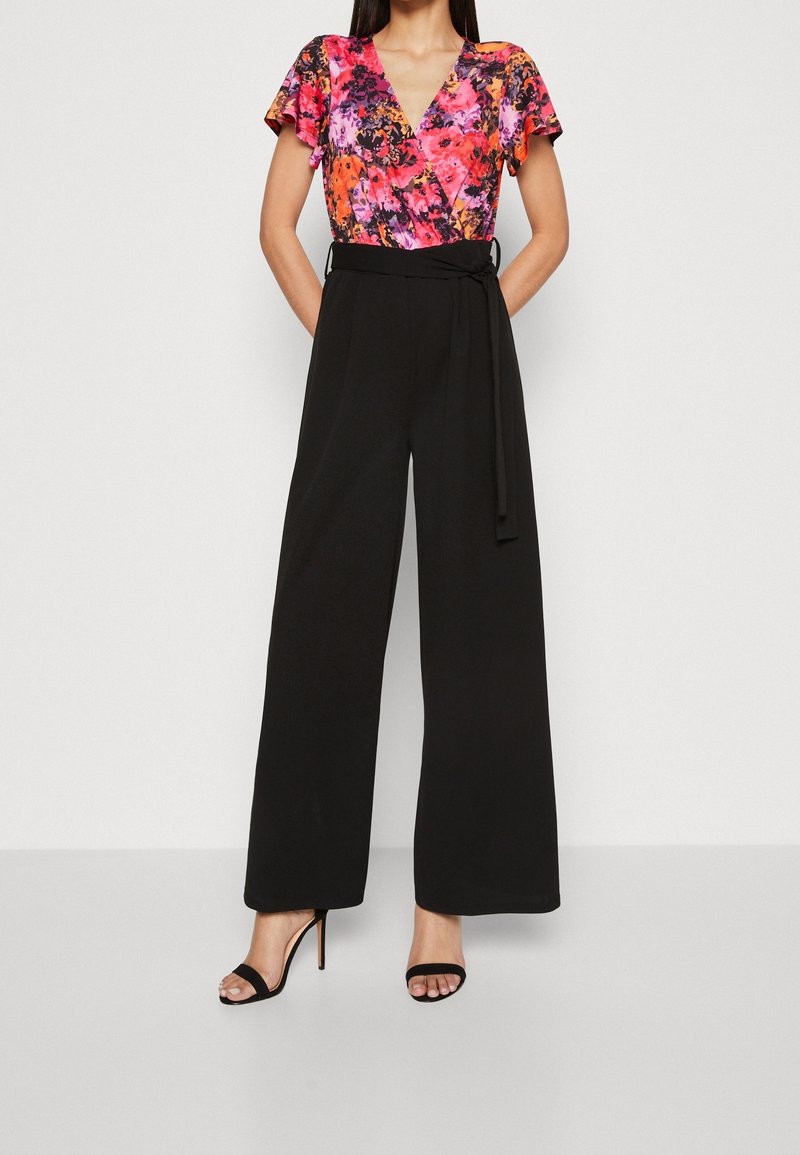 Femme portant un haut cache-cœur floral à manches courtes et un pantalon noir large taille haute avec ceinture à la taille, assortis à des sandales à talons aiguilles noires.