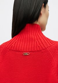 Punainen ribbiknitti neulepaita, jossa on korkea kaulus ja kultainen logo takana. Ominaisuuksiin kuuluu istuva muoto ja sileä tekstuuri.