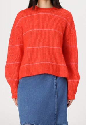 Pull en tricot rayé rouge, avec une coupe décontractée, une texture côtelée et un col rond. Associé à une jupe en jean pour un contraste.