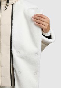 Bílý fleece kabát s texturovaným povrchem, s průhlednými knoflíky a měkkým límcem. Přes béžovou prošívanou bundu.