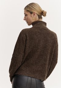 Brauner, strukturierten Pullover mit hohem Kragen, lässiger Passform und gerippten Details an den Ärmeln. Getragen mit schwarzen Hosen.
