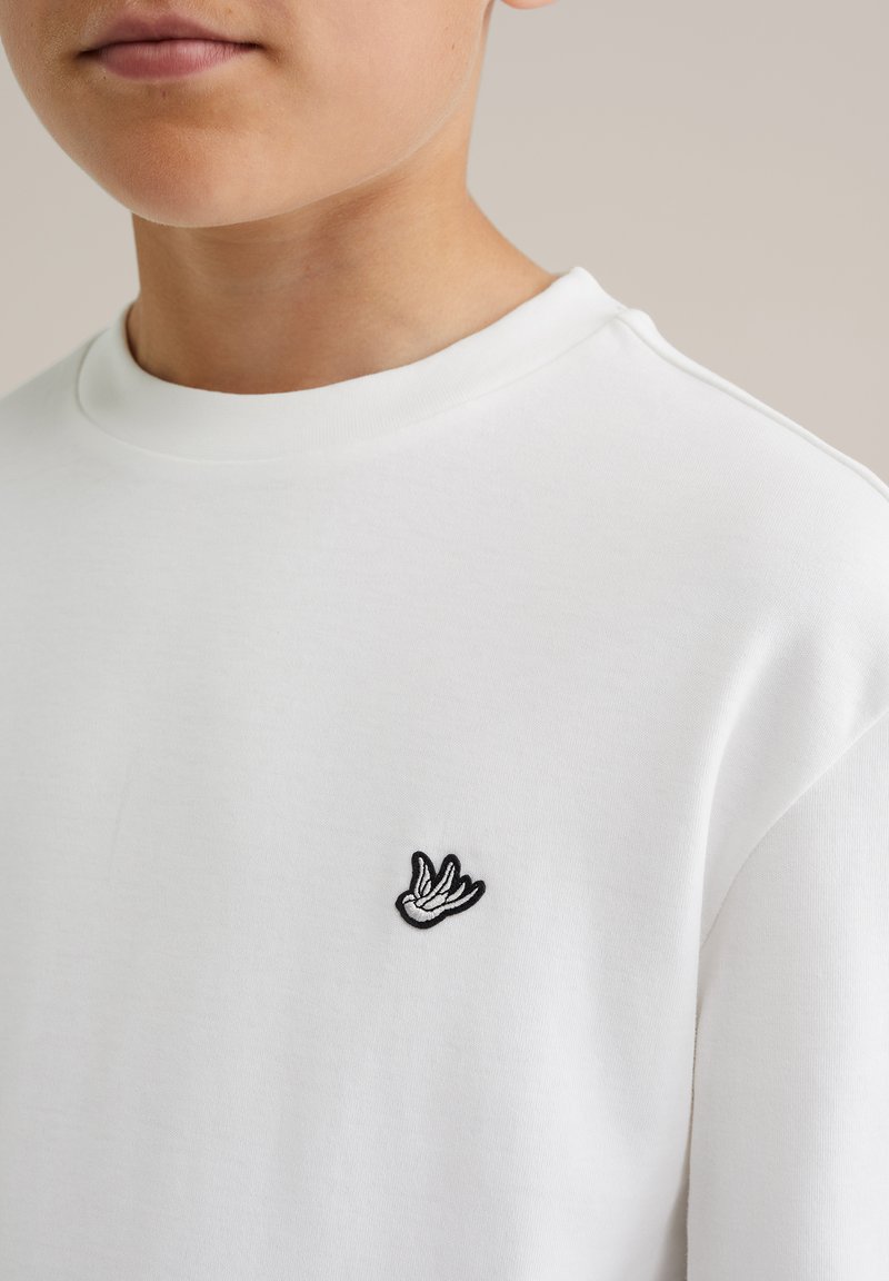 Vit t-shirt med kort ärm som har en svart broderad fågeldesign på vänster bröst, tillverkad av mjuk bomullstyg med en slät struktur.