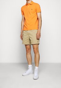 Orange polo-shirt med korta ärmar och krage, tillsammans med beige shorts. Vita hög-top sneakers fulländar looken.