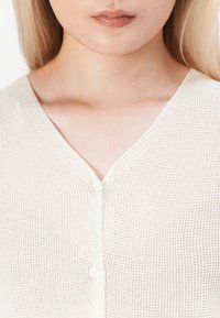 Světle béžový cardigan na knoflíky s texturovaným pleteným vzorem, designem do V a jedním bílým knoflíkem uprostřed přední části.