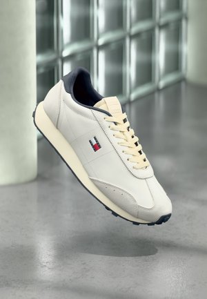 Białe i beżowe sneakersy z granatowym obszyciem, kremowymi sznurówkami i małym logo Tommy Hilfiger po boku, unoszące się nad wypolerowaną betonową podłogą.