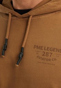 Sudadera con capucha marrón de acabado texturizado. Presenta cordones con puntas negras y una marca impresa en la parte delantera. Diseño informal.