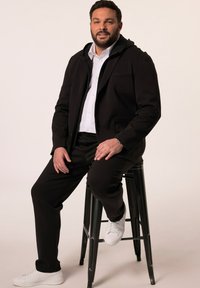 Giacca nera con zip sopra una camicia bianca a maniche lunghe, abbinata a pantaloni neri e sneakers bianche. Il modello è seduto su uno sgabello nero.