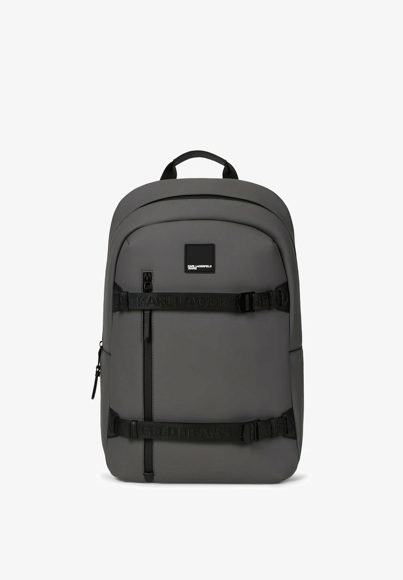 Sac à dos rectangulaire gris foncé avec des bretelles noires réglables sur le devant, une fermeture éclair verticale, une poignée supérieure et un petit patch logo près du centre supérieur.
