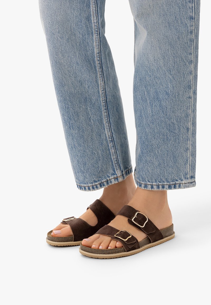 Pieds portant des sandales en daim marron avec deux boucles, associées à un jean denim bleu clair sur fond blanc.