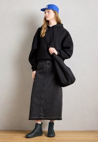 Jeune femme portant une casquette bleue, un sweat à capuche noir surdimensionné, une longue jupe en denim noir, des baskets montantes noires et portant un sac à bandoulière noir.