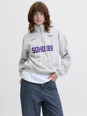 Personne aux cheveux bouclés roux portant un sweat-shirt gris à fermeture éclair au quart avec "SOHO 99" en violet, un t-shirt blanc en dessous, et un jean bleu.