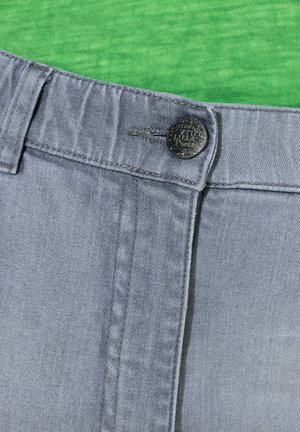 Jeans de mezclilla gris con cierre de botones, que presentan una textura suave y detalles de costura sutiles, sobre un fondo de camisa verde.
