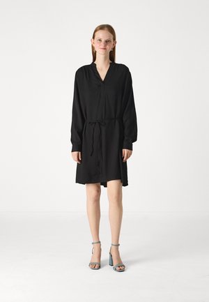 Abito nero con maniche lunghe, scollo a V e vita con laccetto. Tessuto morbido e design a lunghezza ginocchio abbinato a sandali con tacco leggeri blu chiaro.