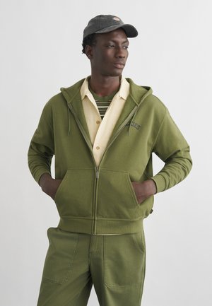 Ung mann iført olivengrønn hettejakke med glidelås, matchende bukser, beige skjorte med knapper, stripete t-skjorte og mørk caps, stående med hendene i lommene.