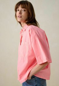 Oversized, short-sleeve shirt in bright pink cotton, featuring a button placket and patch pocket, paired with blue denim jeans.