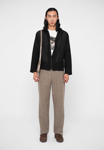 Giacca nera a costine con cerniera frontale, indossata sopra una t-shirt bianca con grafica, abbinata a pantaloni chiari in tessuto strutturato e scarpe marroni.