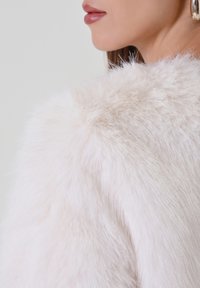 Kleidungsstück aus Kunstpelz in sanftem Weiß, das eine flauschige Textur zeigt. Mit abgerundetem Schulterdesign, wobei glatte Hauttöne daneben sichtbar sind.