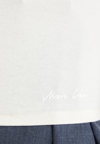 Weißes Baumwoll-T-Shirt mit glatter Textur. Verfügt über einen abgerundeten Saum und ein gesticktes Logo in Laufschrift in der unteren linken Ecke.