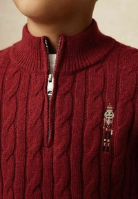 Punane koonuskeermest kudumiga kampsun kõrge krae ja pooltõmblukuga; vasakul rinnal on väike tikitud pähklimurdja kujutis.