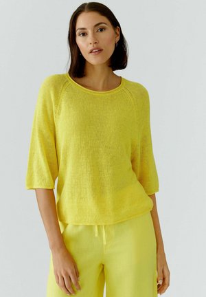 Vrouw met donker schouderlang haar, die een losse gele gebreide trui met ellebooglange mouwen en bijpassende gele broek draagt.