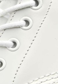 Primo piano di una sneaker in pelle bianca, con lacci bianchi intrecciati, occhielli in metallo, cuciture visibili e una superficie liscia e testurizzata.