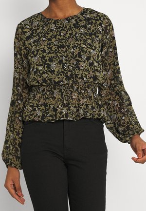 Femme portant un chemisier à manches longues vert et noir à motifs floraux avec une taille cintrée et un pantalon noir sur un fond uni.