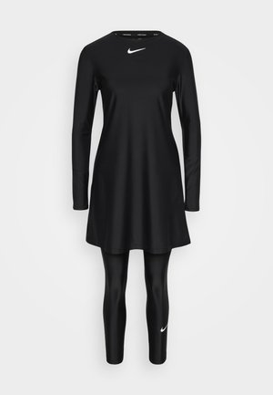 Černé dlouhé sportovní šaty s kulatým výstřihem, s bílým logem Nike na hrudi, spárované s černými legínami. Hladká textura.