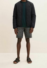 Giacca bomber nera trapuntata con colletto rotondo, abbinata a un maglione verde scuro, pantaloni corti di denim grigi e sandali neri.