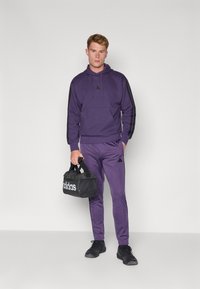 Sweatshirt roxo com detalhes e logótipo pretos, combinado com calças roxas a condizer. A segurar um saco de desporto preto com logótipo branco. Ténis pretos.
