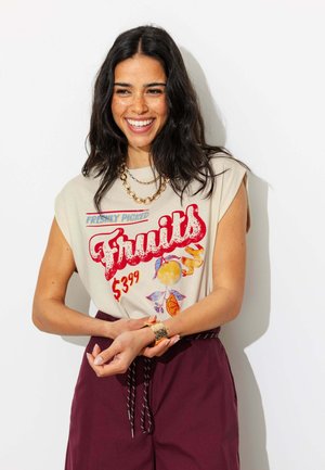 Jeune femme aux longs cheveux foncés souriante, portant un débardeur avec le texte « Fruits fraîchement cueillis 3,99 $ » et un pantalon grenat.