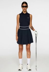 Laivastonsininen hihaton golfmekko, jossa on pliseerattu hameosa ja valkoiset yksityiskohdat. Päällä valkoiset sukat ja tennarit, kädessä golfmaila. Aurinkolasit päässä.