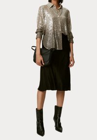 Vrouw in een zilveren pailletten blouse met knopen, zwarte midi-rok, zwarte enkellaarsjes en met een zwarte handtas voor een witte achtergrond.