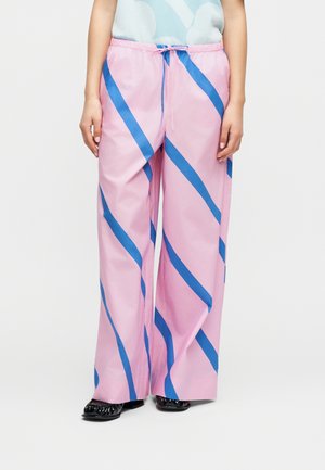 Person iført løstsiddende lyserøde bukser med brede blå diagonale striber og sorte sko, stående mod en ensfarvet hvid baggrund.