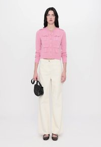 Cardigan en tricot rose clair avec poches avant, associé à un pantalon large crème. Le mannequin tient un petit sac à main noir, se tenant sur un fond neutre.