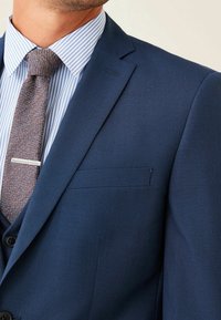Marineblauw pak met een gestructureerde afwerking, gecombineerd met een lichtblauw gestreept overhemdd en een bruine herringbone stropdas met zilveren accenten.