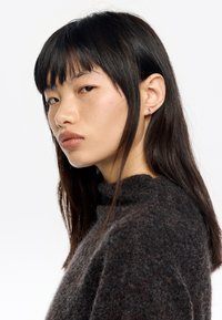 Maglione a maglia grigio scuro con colletto alto, indossato da una persona con capelli neri lisci e orecchini sottili. Tessuto liscio, design minimale.