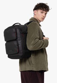 Mochila negra con forma estructurada, dos bolsillos frontales, detalles rojos y tirantes ajustables, llevada en la espalda de un modelo con una chaqueta de forro polar verde.