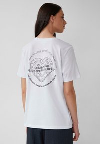 T-shirt en coton blanc avec un design de cœur noir imprimé au dos, entouré de texte. Coupe ample, manches courtes et col rond.
