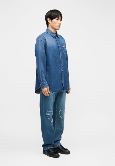 Jeune homme debout portant une chemise en denim bleu et un jean avec des patchs en forme de cœur usés sur les genoux, baskets noires, fond neutre.