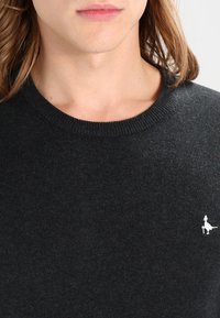 Ciemnoszary sweter z okrągłym dekoltem i prążkowanymi mankietami. Na lewej piersi małe białe logo. Wykonany z miękkiej dzianiny.