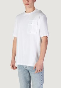 T-shirt bianco in cotone con collo tondo e maniche corte, dotato di un taschino sul lato sinistro del petto. Abbinato a jeans azzurri chiari.