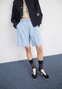 Shorts en denim bleu clair avec des plis, portés avec une veste noire et des chaussettes noires, associés à des baskets foncées avec des accents blancs.